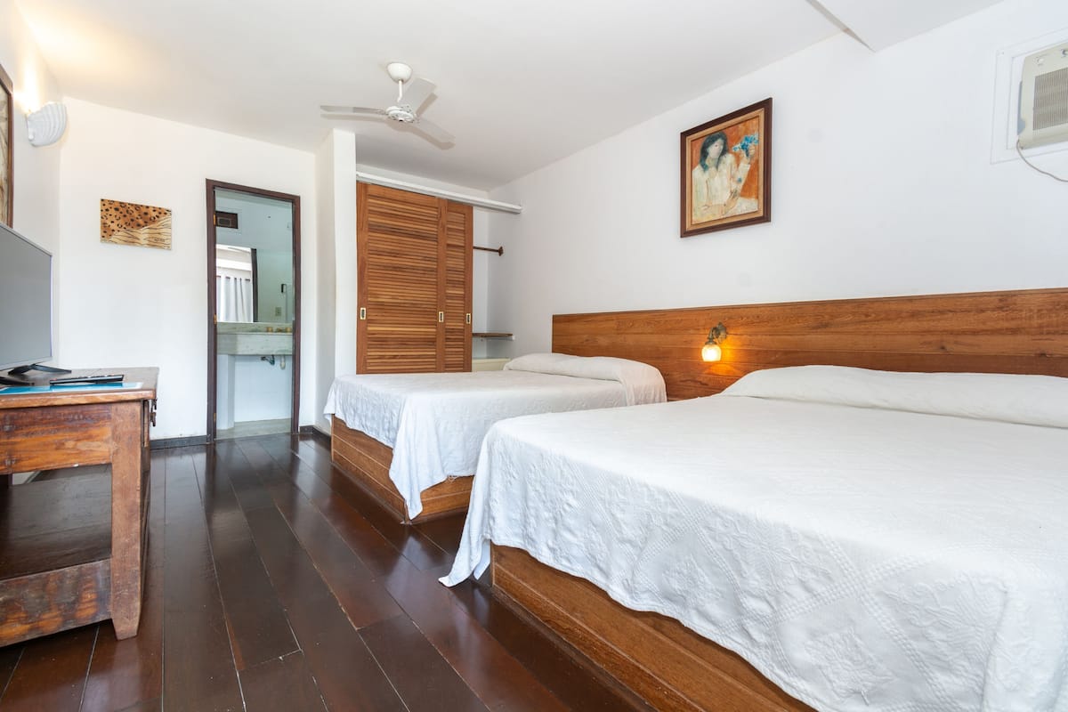 Une chambre d’hôtel avec deux lits simples, un bureau en bois, un ventilateur de plafond et un tableau au mur.