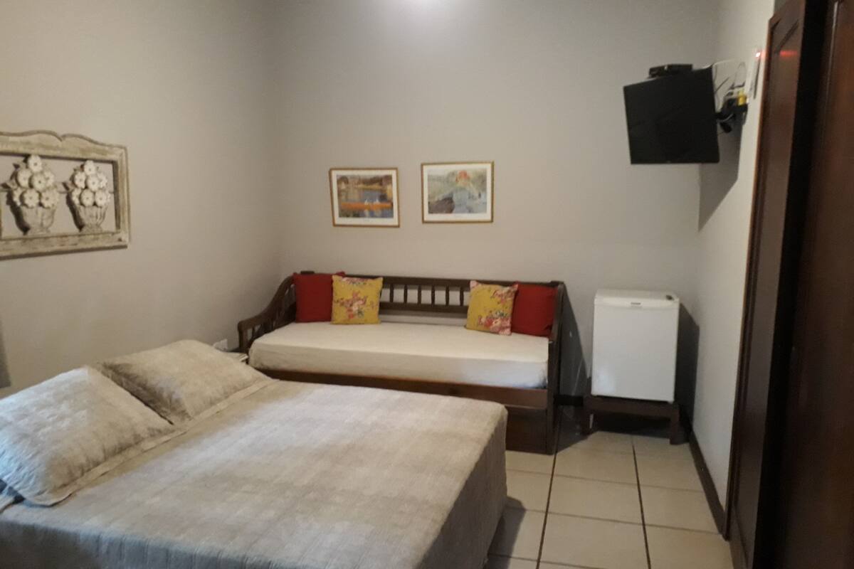 A bedroom with a bed, a small bedside table, a wall-mounted heater, and two framed pictures.