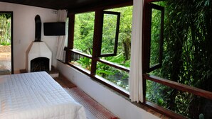 A bedroom with a bed, a fireplace, a TV, and large windows with a view of greenery. - Hotel Daniela (Itatiaia)