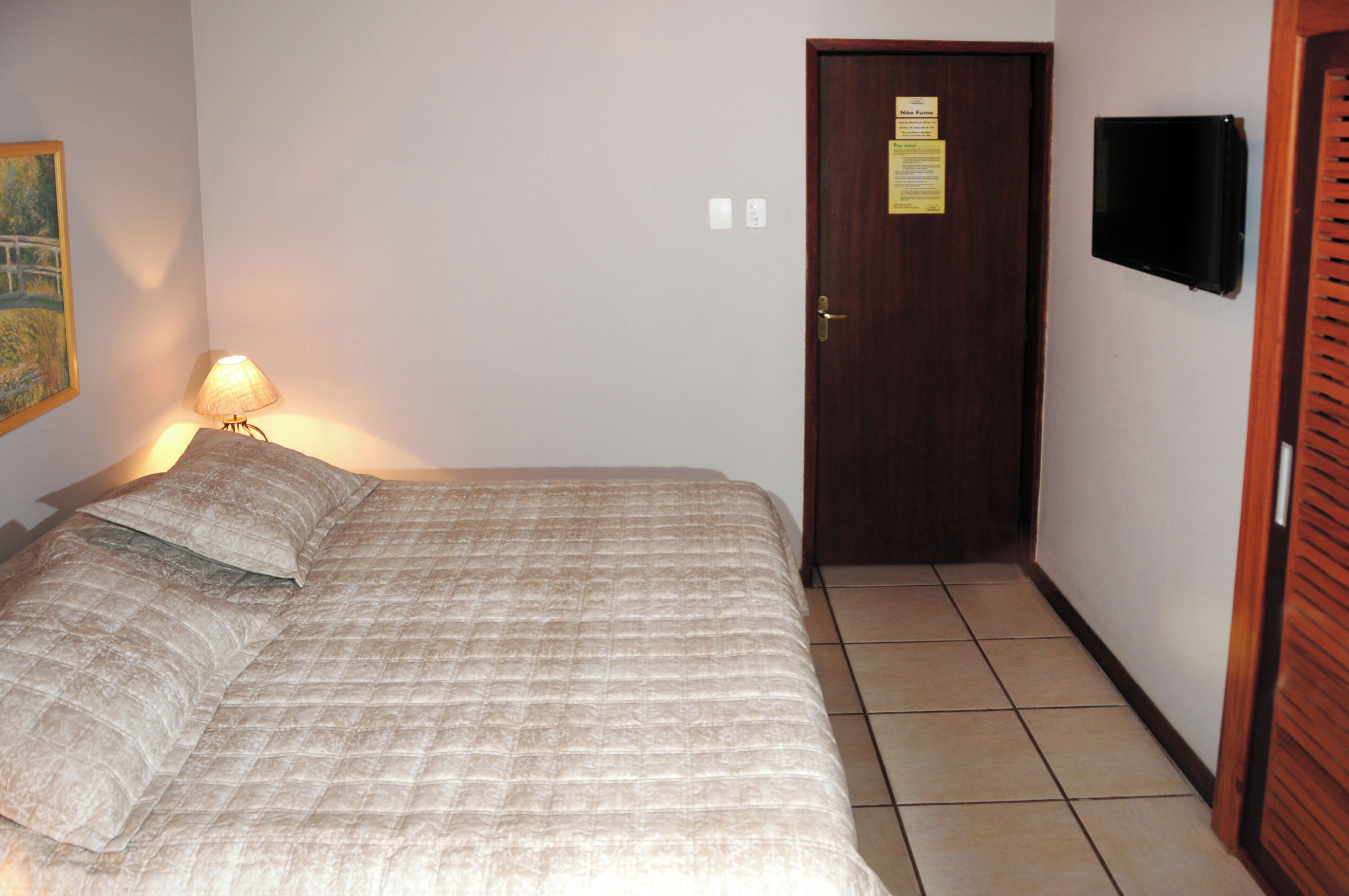 A bedroom with a bed, a lamp, a door, and a wall-mounted TV.