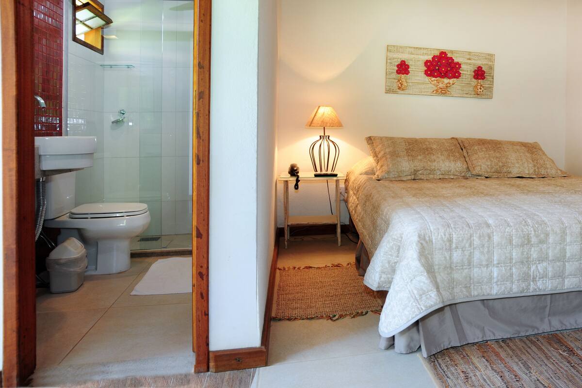 A bedroom with a bed, a bedside table with a lamp, a wall-mounted painting, and a bathroom with a toilet and sink.