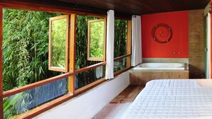 A bedroom with a large bed, a window with a view of bamboo, a bathtub, and a red wall. - Hotel Daniela (Itatiaia)