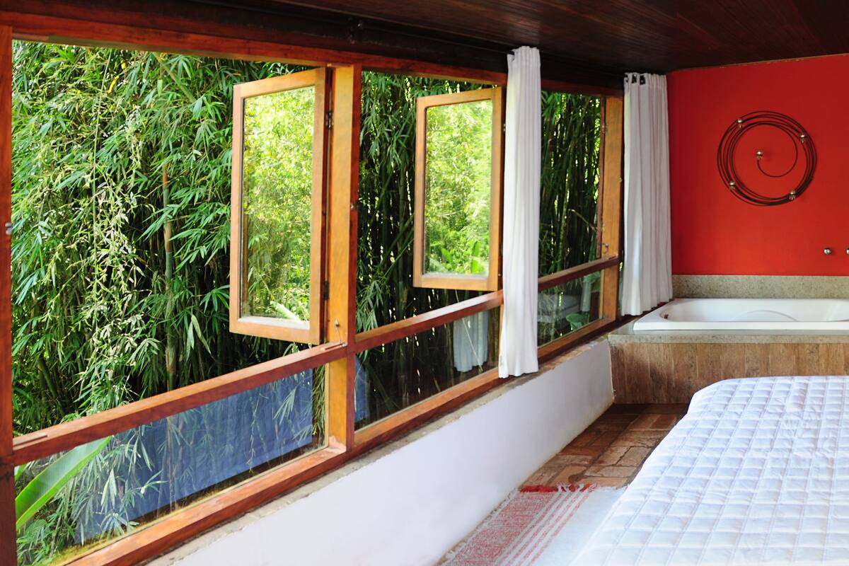 A bedroom with a large bed, a window with a view of bamboo, a bathtub, and a red wall.
