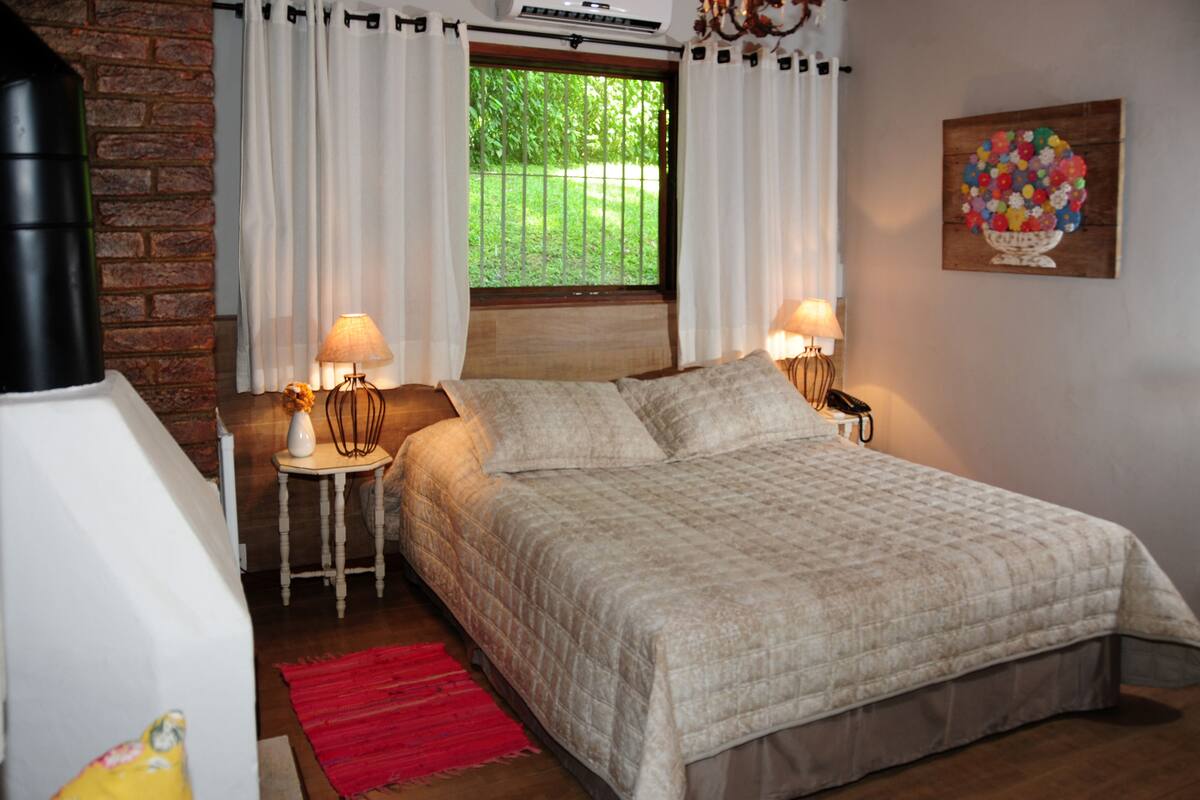 A bedroom with a bed, two bedside lamps, a window with curtains, a radiator, a red rug, and a wall-mounted painting.
