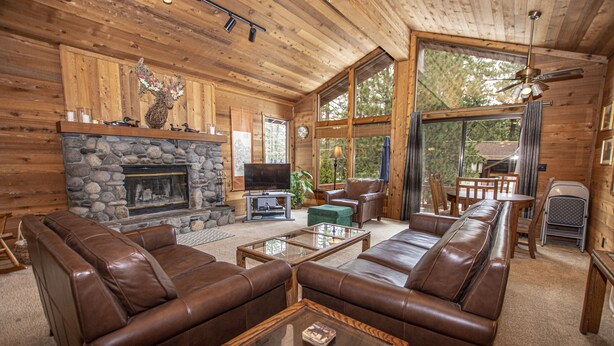 A cozy cabin interior with a stone fireplace, leather sofas, a television, and large windows offering a view of the outdoors. - Vance by Hauserman Rentals (Tahoe City)