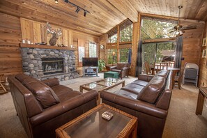 A cozy cabin interior with a stone fireplace, leather sofas, a television, and large windows offering a view of the outdoors.