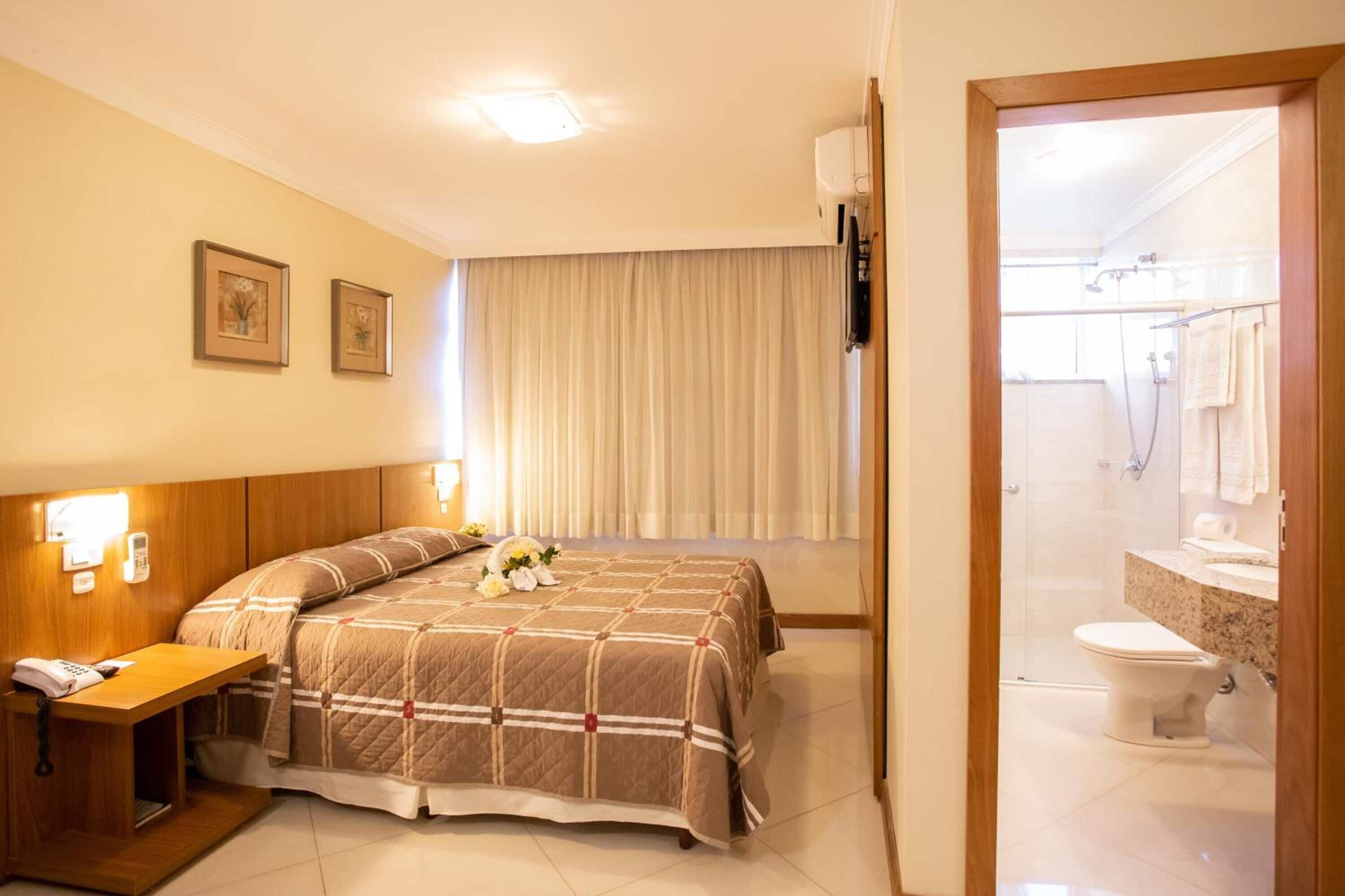 Habitación de hotel con cama, mesita de noche, lámpara y vista al baño.