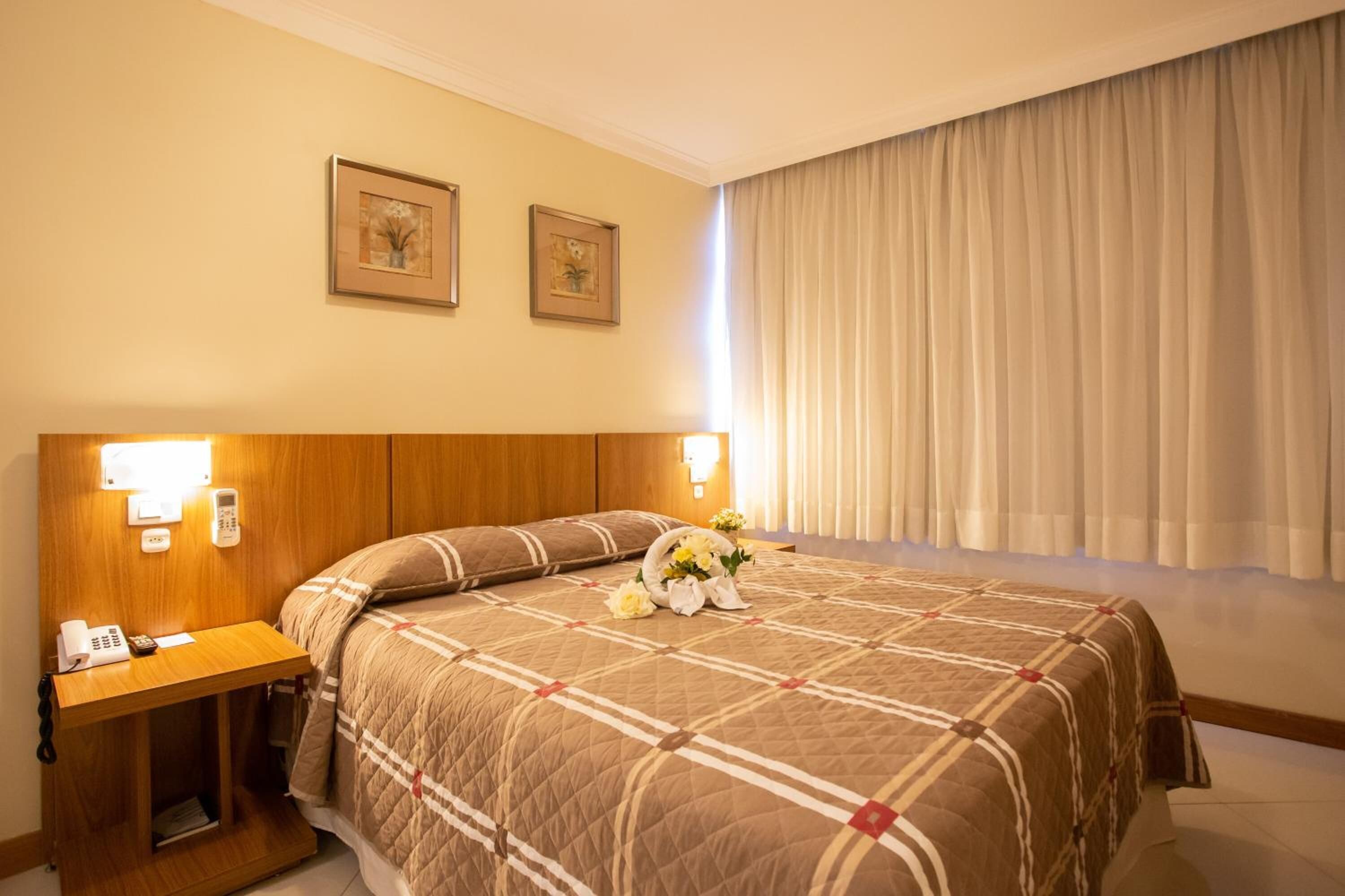 A hotel room with a large bed, a wooden headboard, a bedside table with a phone and lamp, and two framed pictures on the wall.