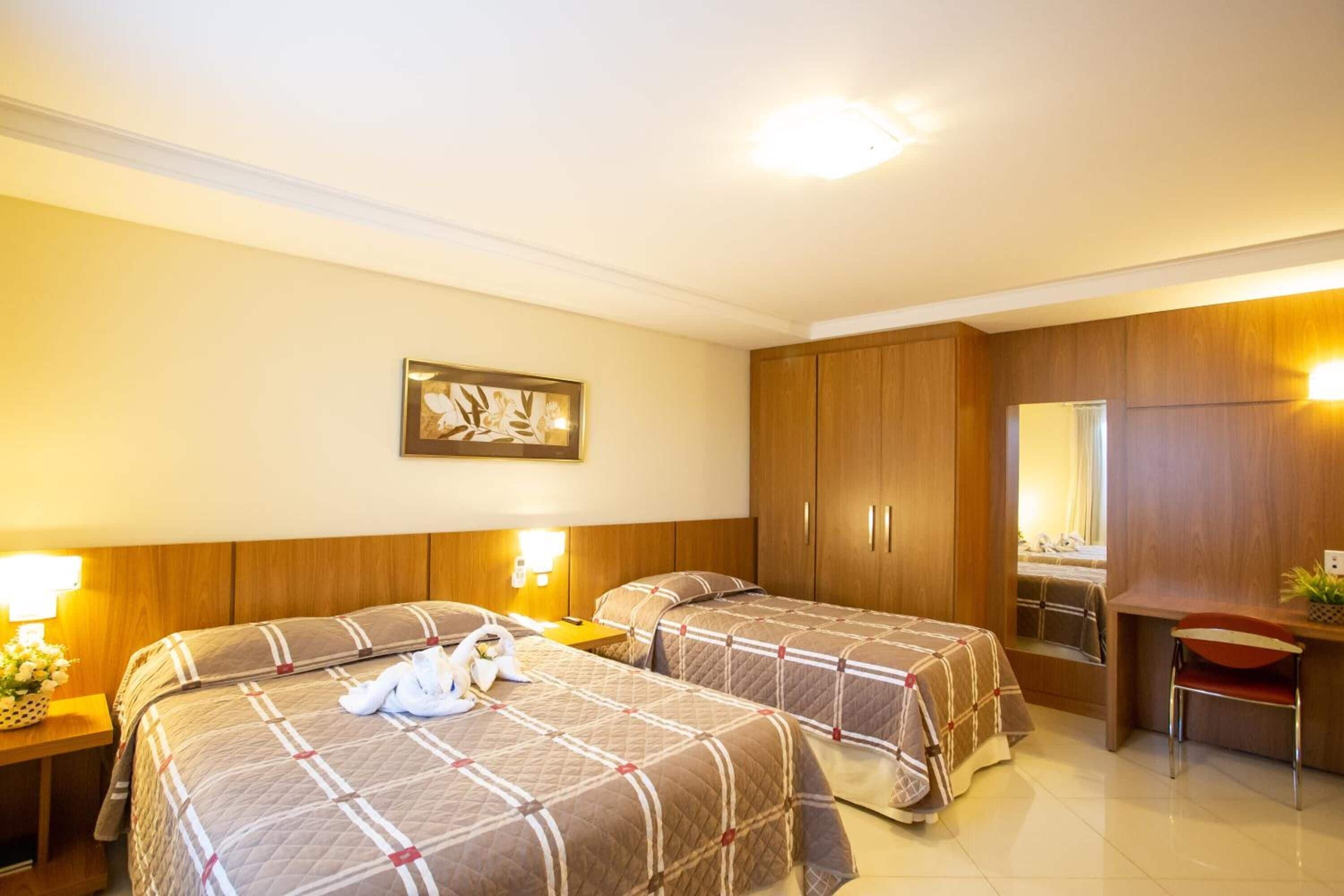 A hotel room with two beds, a desk, and a chair. There is a framed picture on the wall and a lamp on the nightstand.