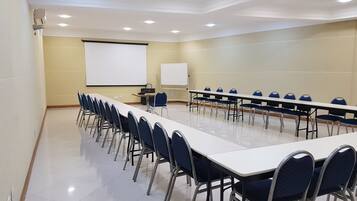Uma sala de conferências com uma mesa comprida, fileiras de cadeiras, uma tela de projetor e um quadro branco.