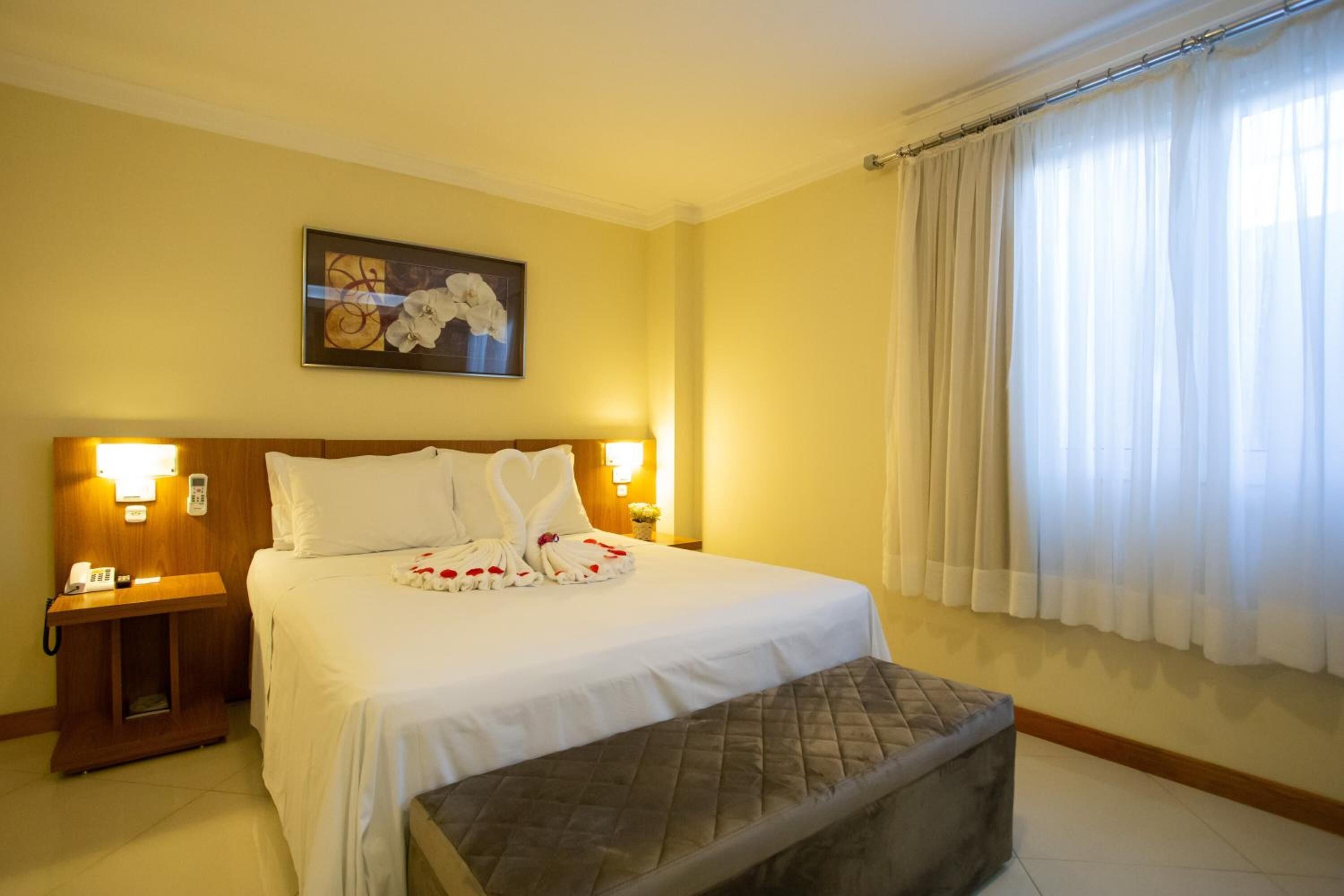 A hotel room with a bed, bedside lamps, a nightstand, a window with curtains, and a painting on the wall.