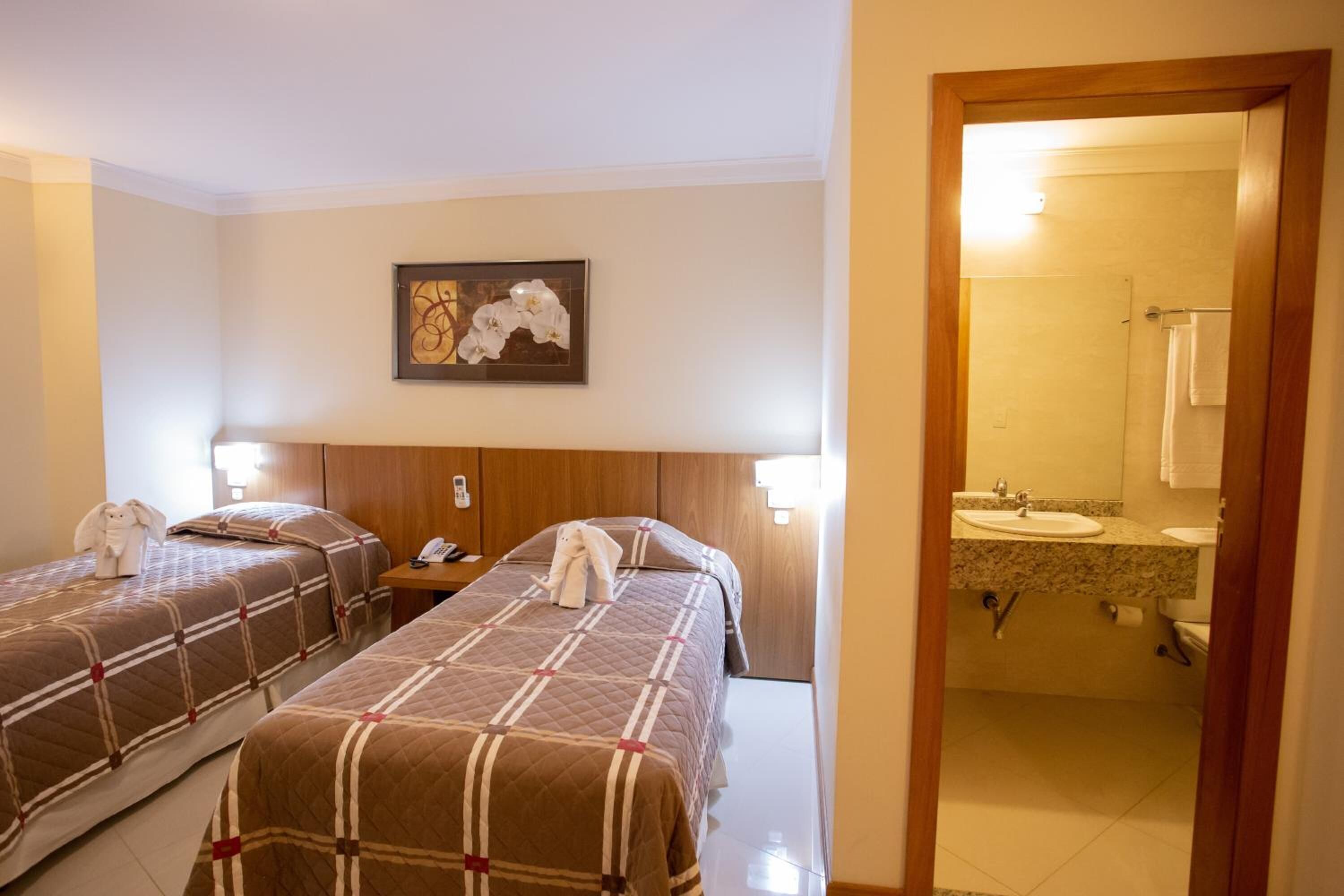 Habitación de hotel con dos camas, cabecero de madera, un cuadro, una mesita de noche, teléfono y un baño visible a través de una puerta abierta.