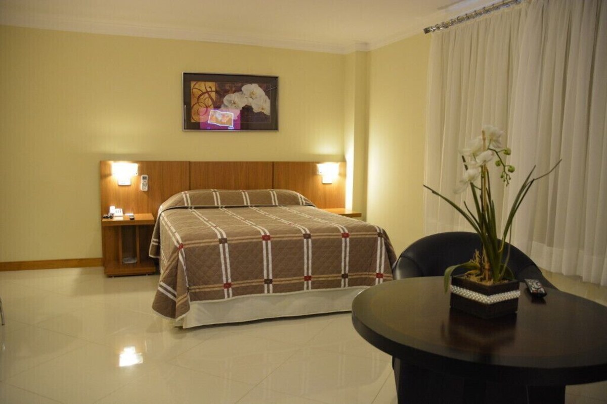 A hotel room with a bed, bedside lamps, a table with a vase of flowers, and a painting on the wall.
