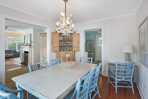 A dining area with a large table, blue chairs, a chandelier, wooden cabinets, and a kitchen in the background.