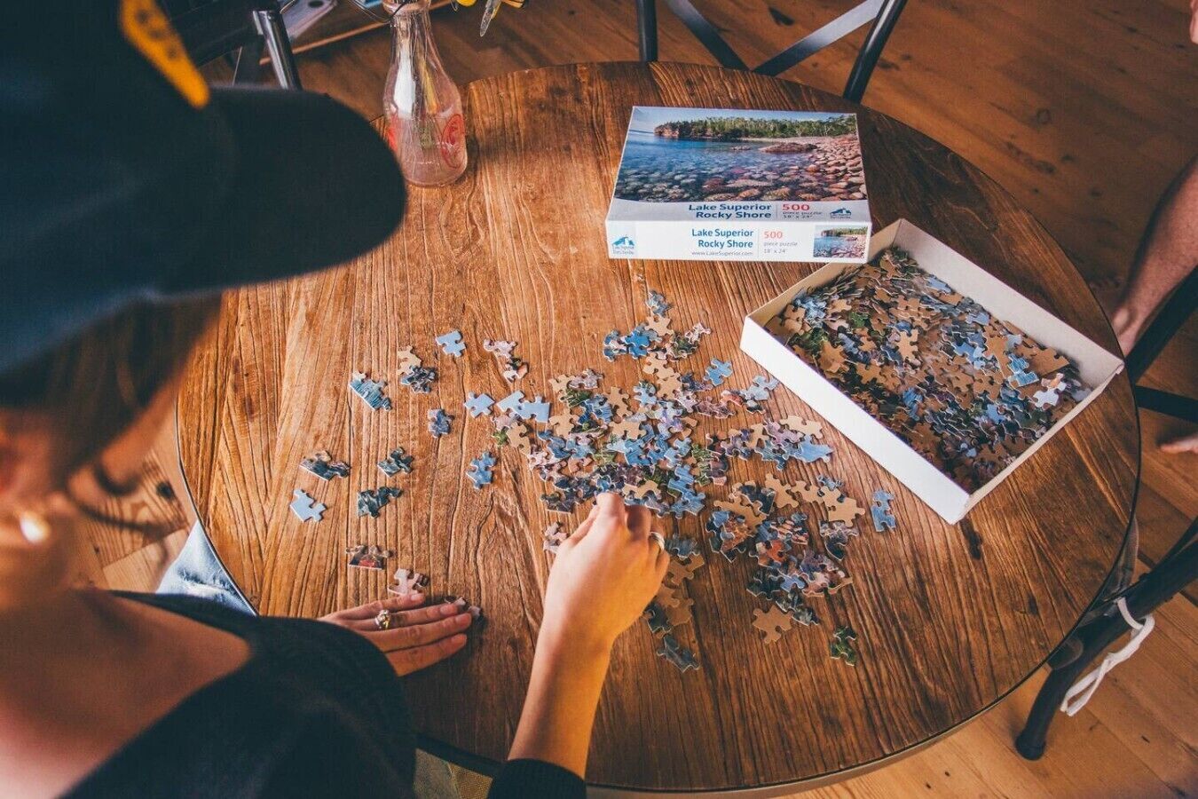 A person assembling a puzzle on a wooden table with a box labeled 'Lake Superior Rocky Shore 500' and puzzle pieces scattered around.