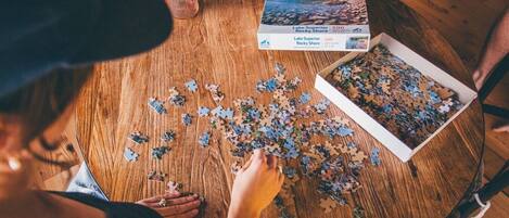 A person assembling a puzzle on a wooden table with a box labeled 'Lake Superior Rocky Shore 500' and puzzle pieces scattered around.