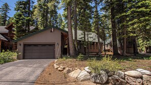 Une maison avec un garage, entourée d’une zone boisée avec de grands arbres et une allée en gravier.
