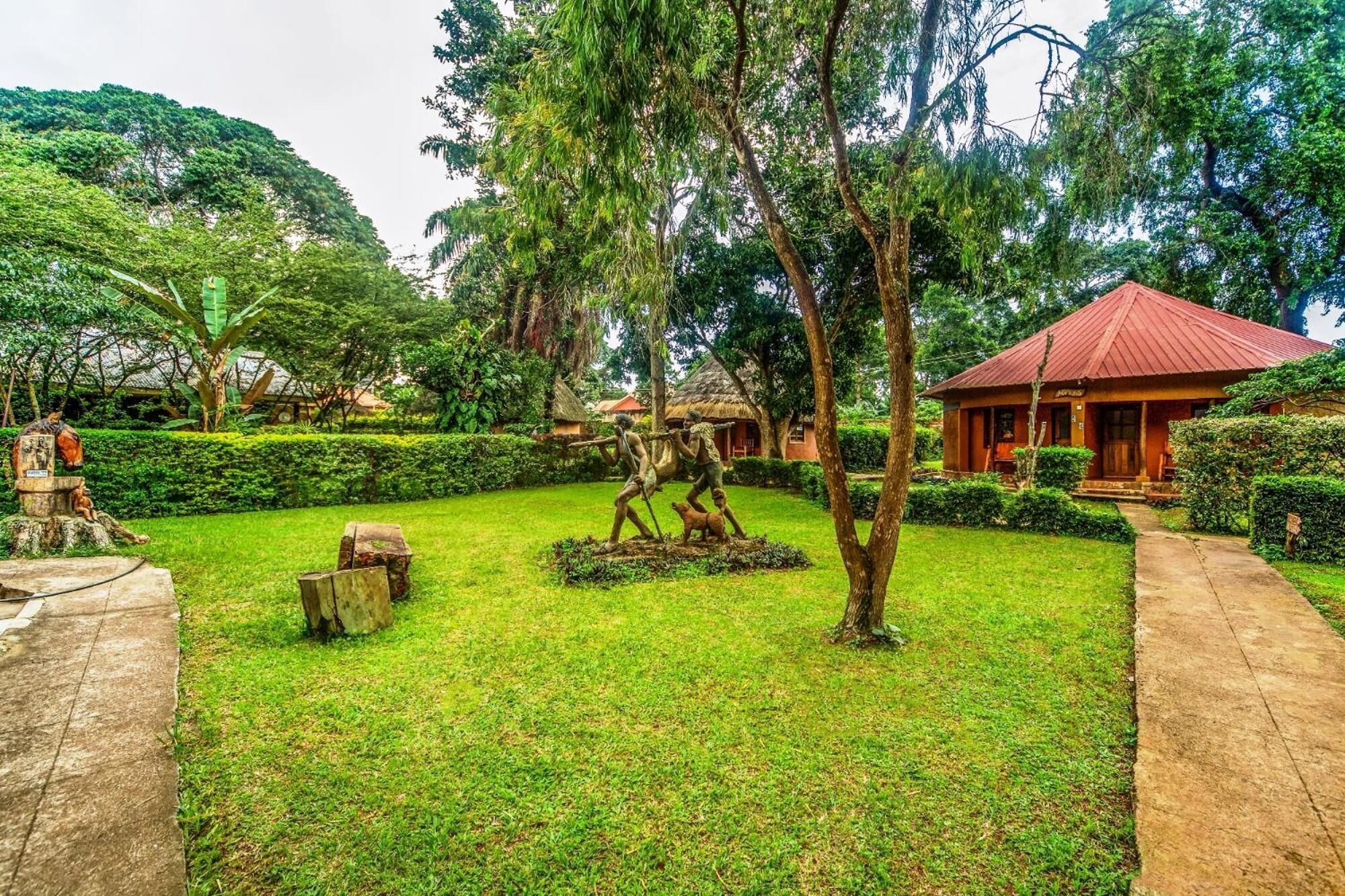 A lush garden with a red-roofed building, a statue of a horse, and a thatched-roof structure.