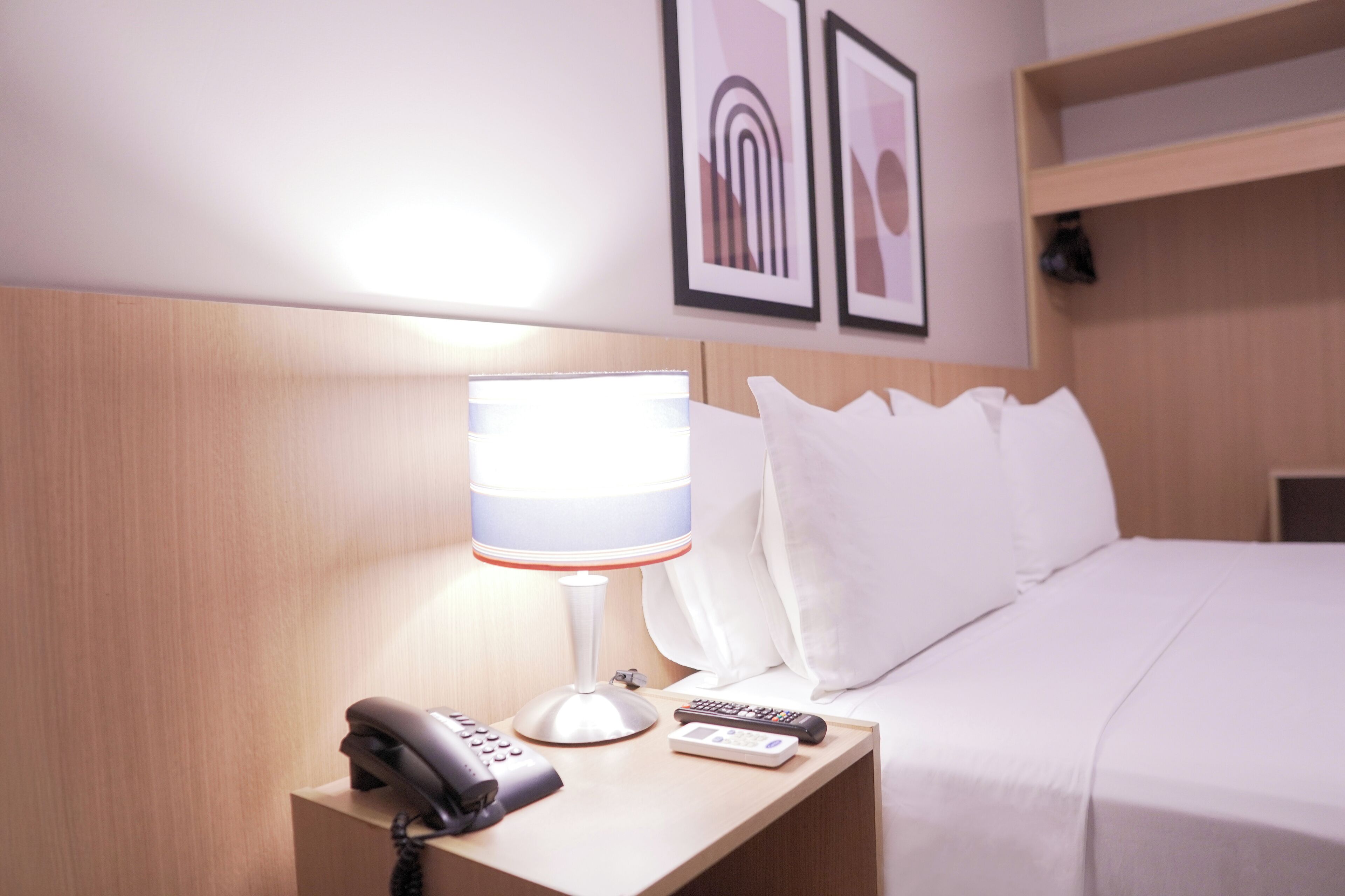 A hotel room with a bed, a bedside table with a lamp, a phone, and a remote control. There are framed pictures on the wall above the bed.