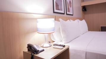 A hotel room with a bed, a bedside table with a lamp, a phone, and a remote control. There are framed pictures on the wall above the bed.