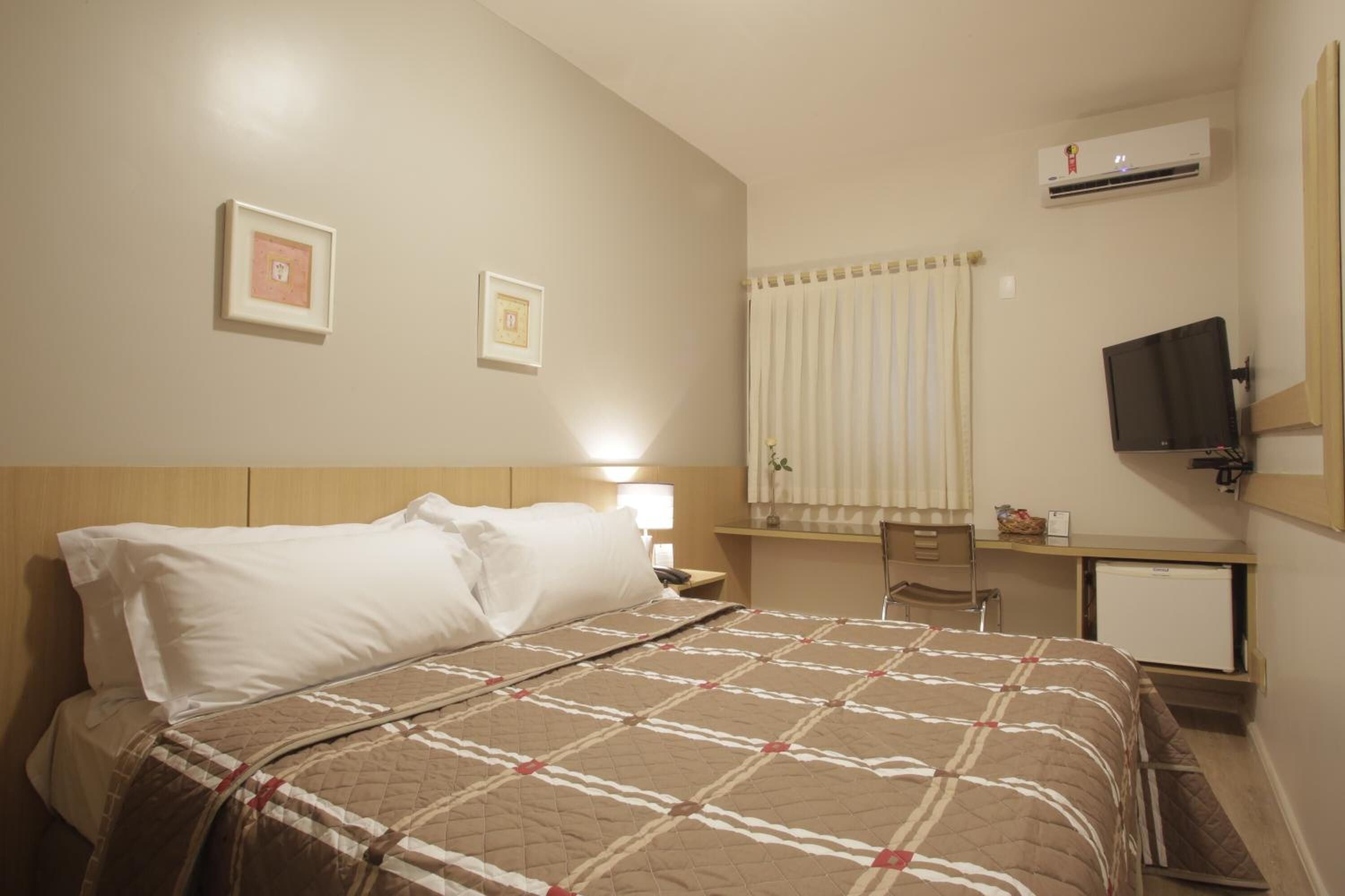 A hotel room with a bed, a desk, a chair, a television, and an air conditioning unit.