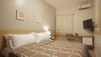 A hotel room with a bed, a desk, a chair, a television, and an air conditioning unit.