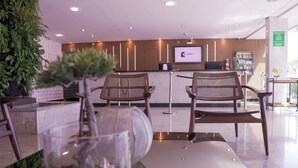 Un área de recepción con un mostrador de madera, dos sillas de mimbre, una mesa de vidrio y una planta grande en maceta.