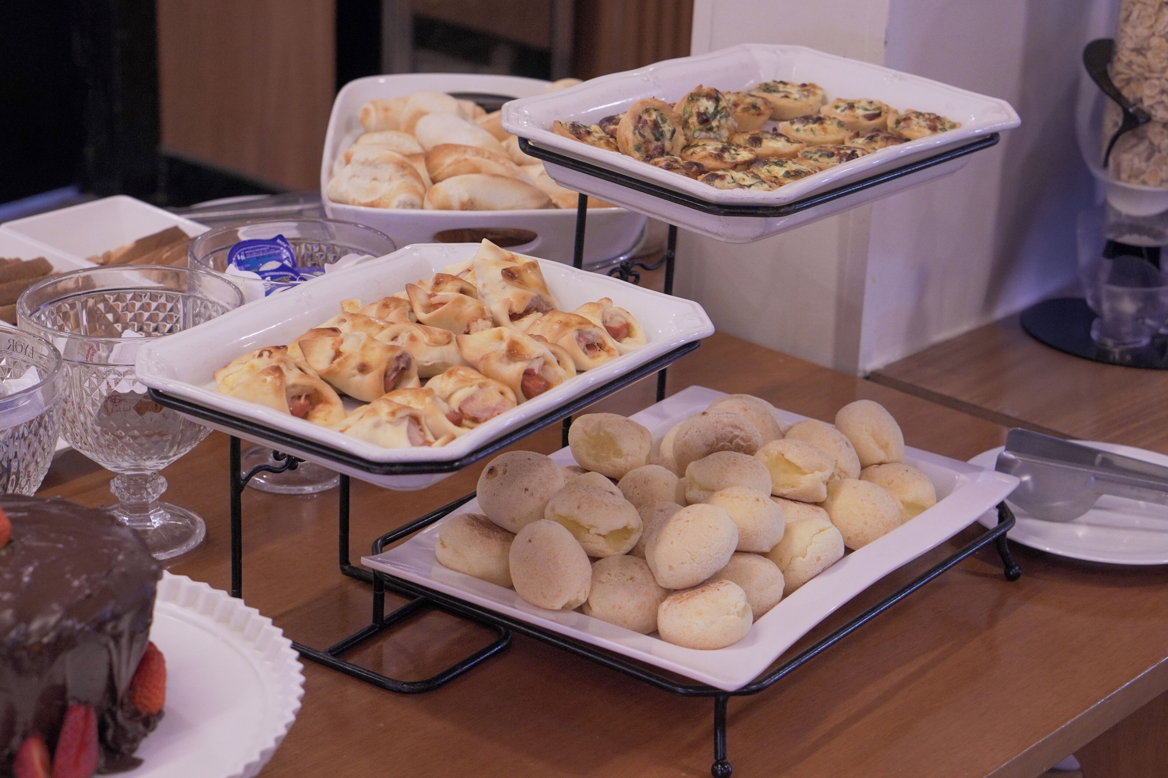 Uma mesa de buffet com diversos alimentos, incluindo pães, doces e uma sobremesa de chocolate.
