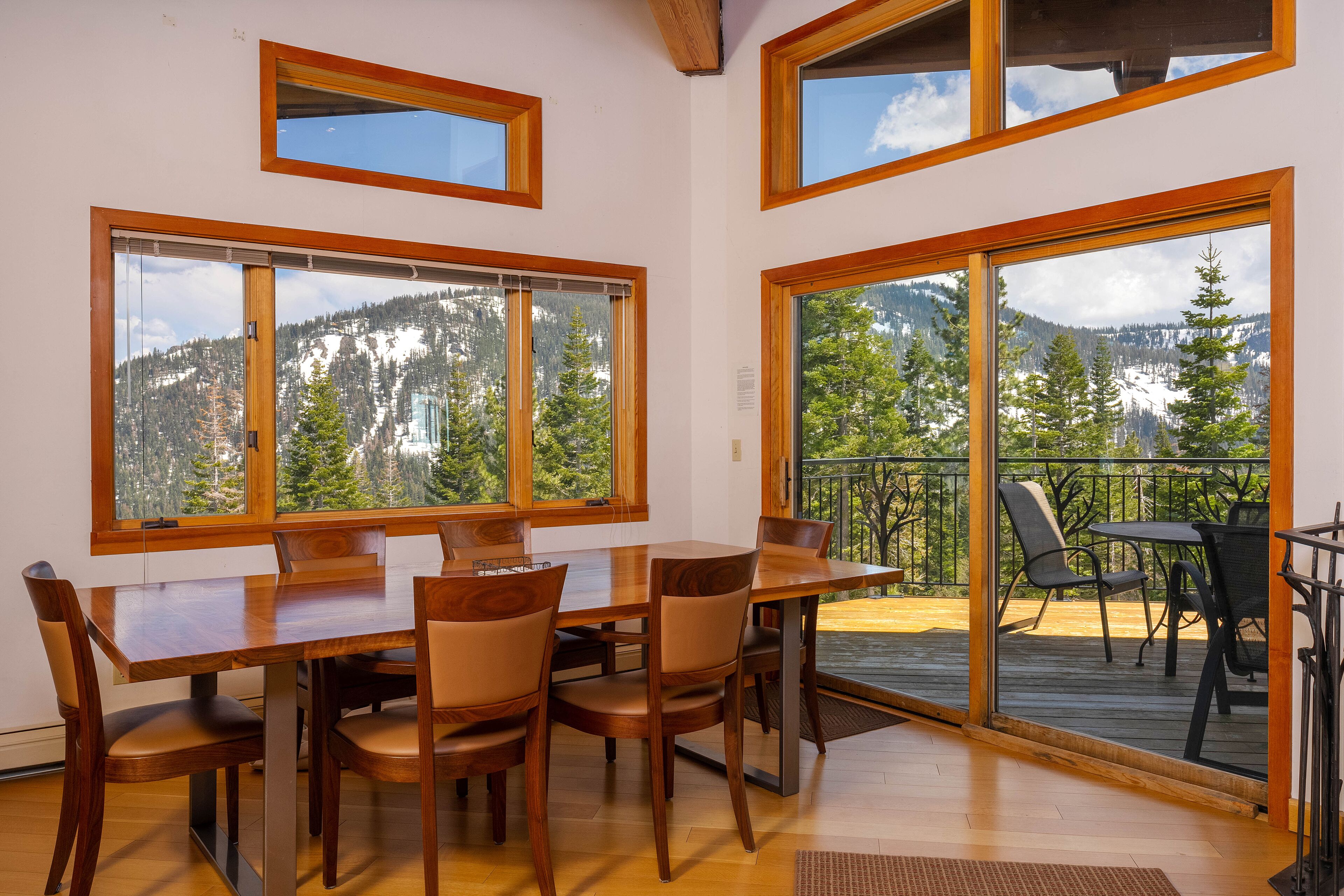 Un espace repas comprenant une table et des chaises en bois, de grandes fenêtres offrant une vue sur les montagnes et un balcon avec des sièges d’extérieur.