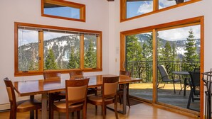 A dining area with a wooden table and chairs, large windows with mountain views, and a balcony with outdoor seating.