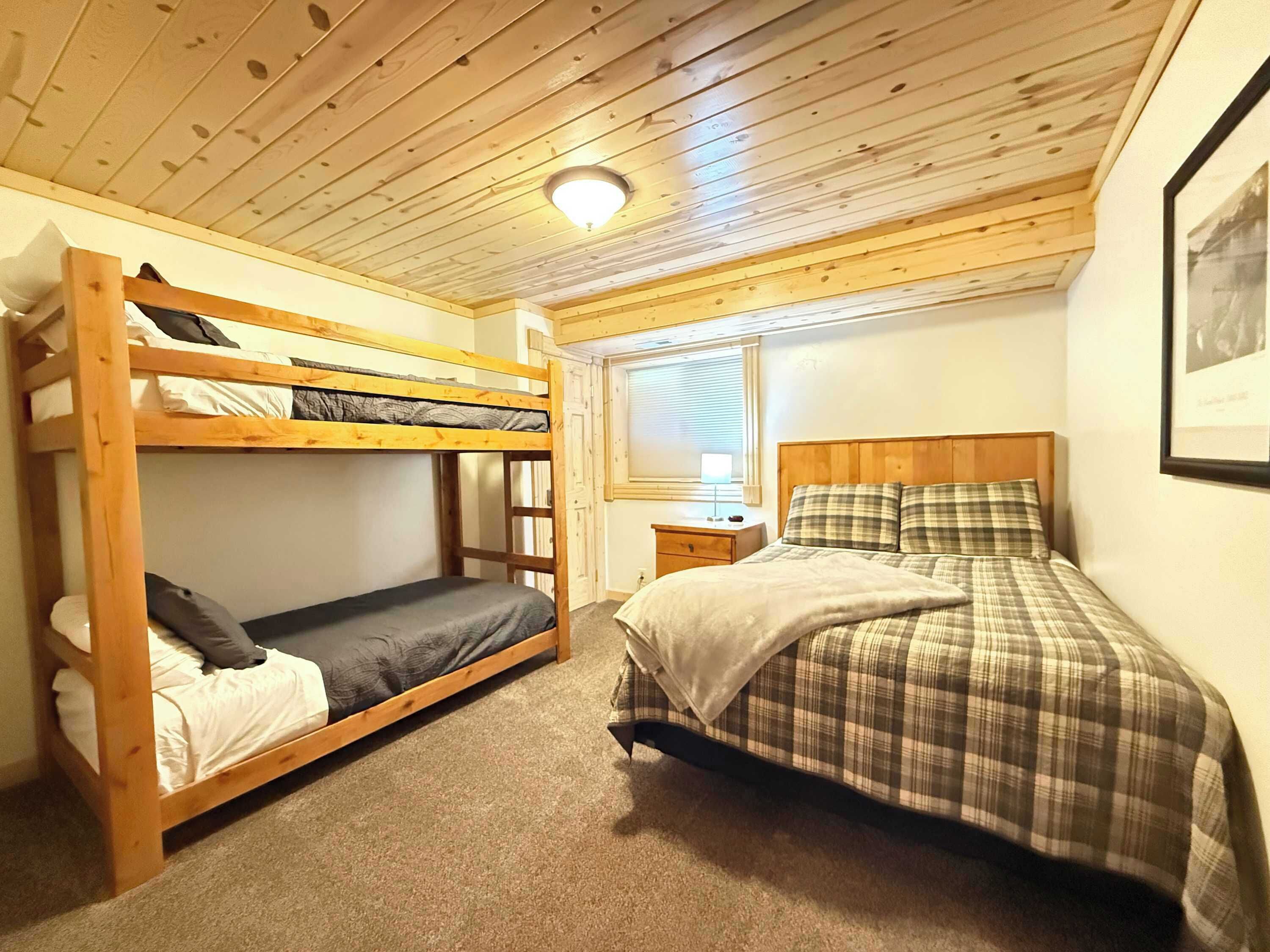 A bunk bed room with a wooden ceiling, a single bed with plaid bedding, a bedside table, and a window with blinds.