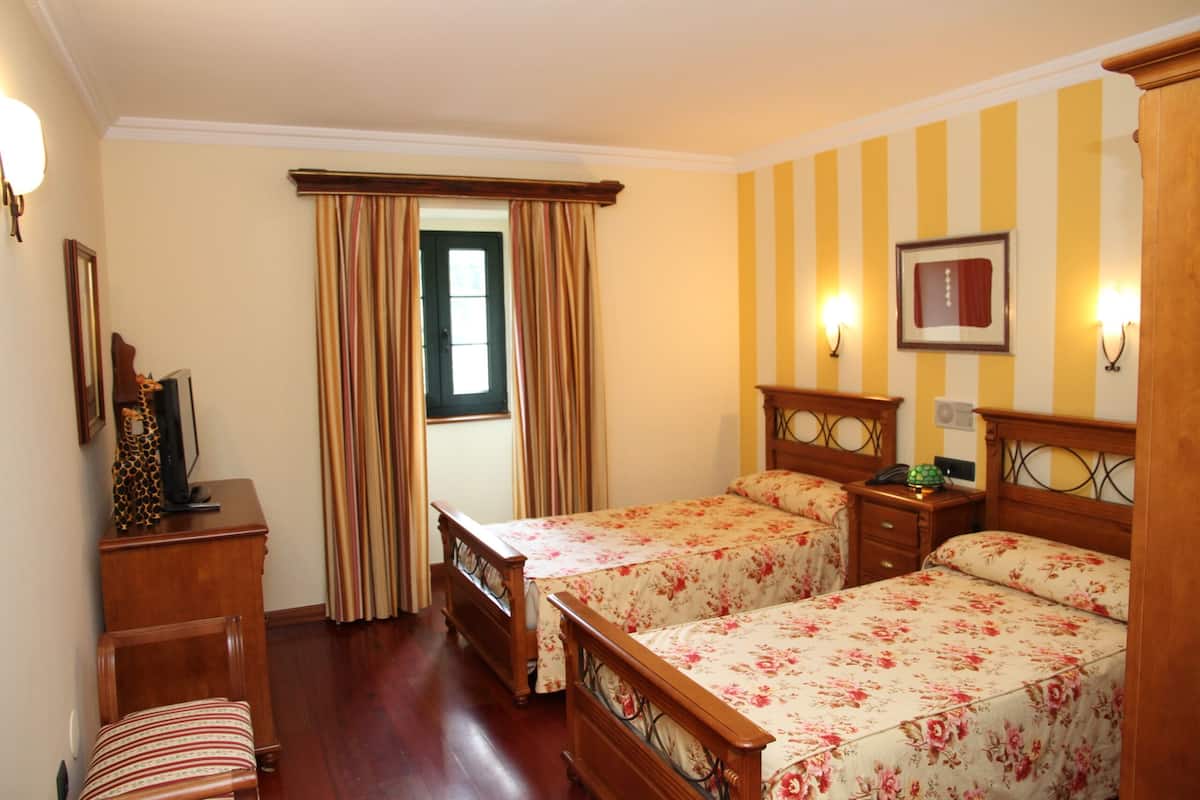 Een hotelkamer met twee houten bedden, een bureau, een stoel, een raam met gordijnen en wandlampen.