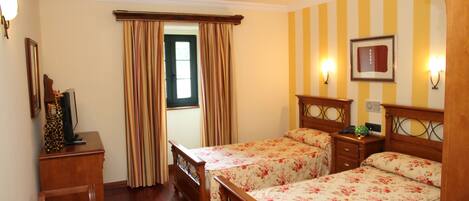 A hotel room with two wooden beds, a desk, a chair, a window with curtains, and wall-mounted lights.