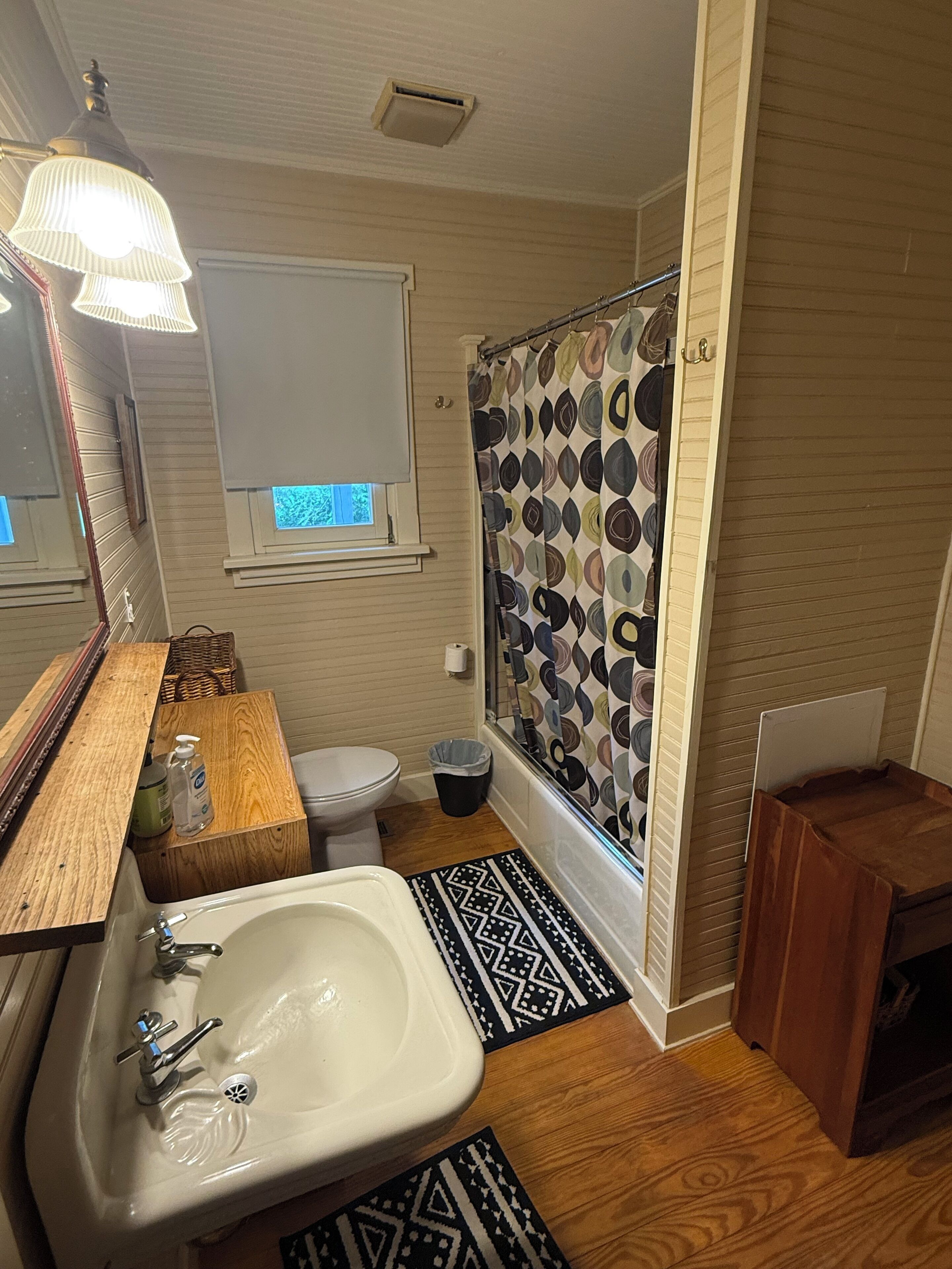 Een badkamer met een houten wastafelmeubel, een witte wasbak, een toilet, een douche met opvallende gordijnen en een klein houten krukje.