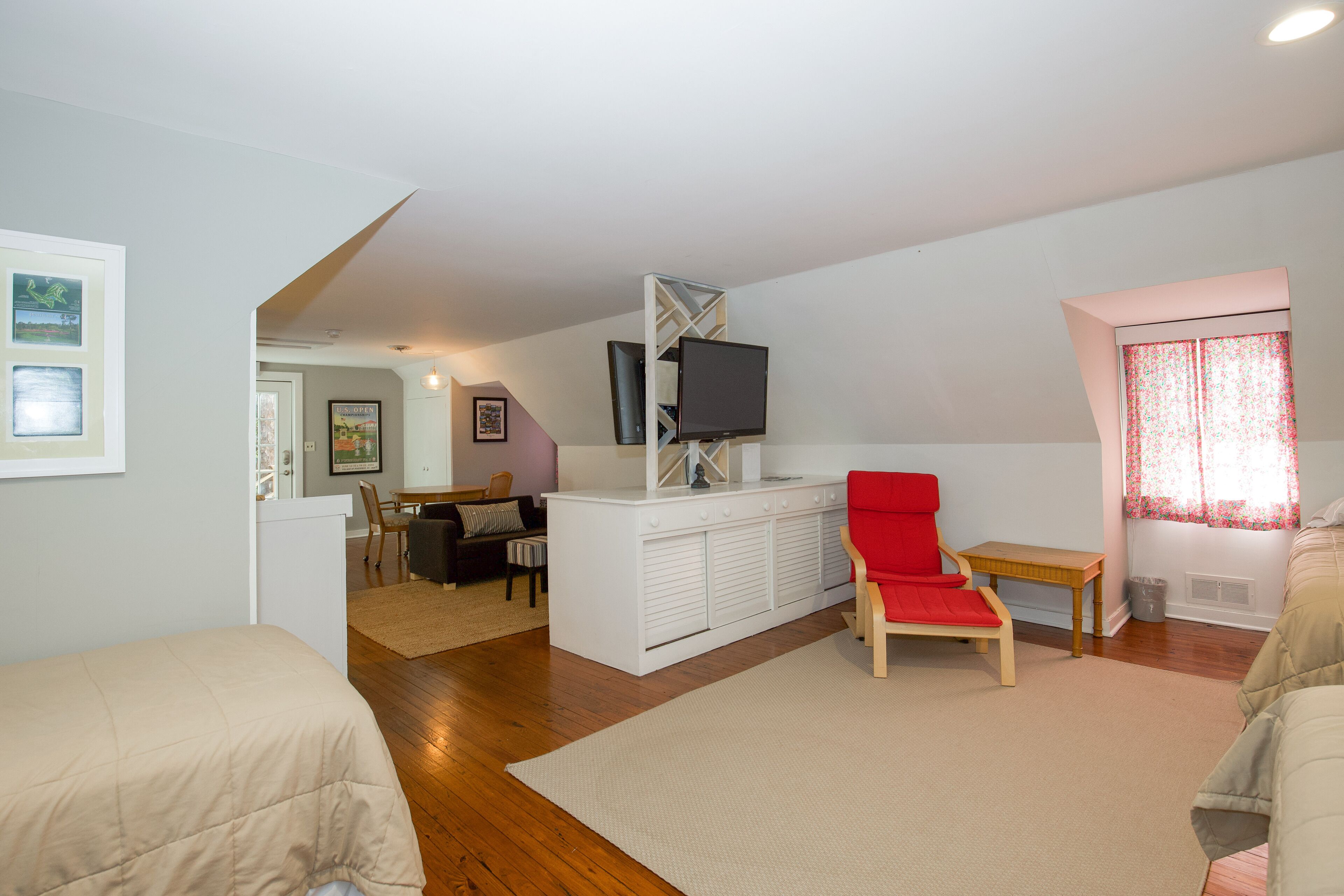 Una habitación con cama, una silla roja, un televisor sobre un mueble blanco y una ventana con cortinas florales.