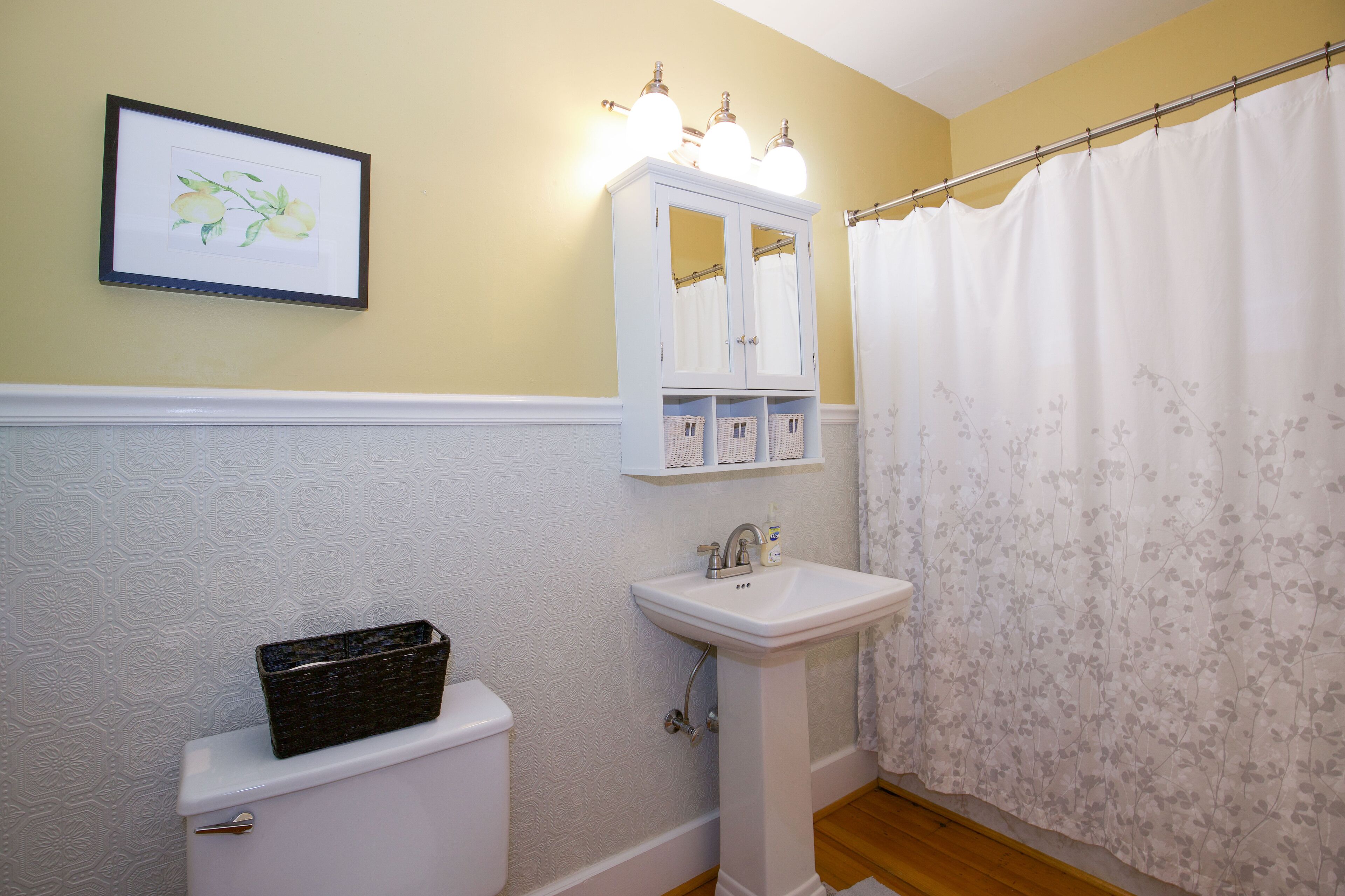 A bathroom with a white toilet, a pedestal sink, a framed floral artwork on the wall, and a shower curtain with a floral pattern.