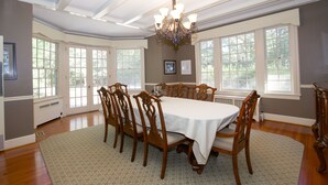 A dining room with a large table, six chairs, a chandelier, and large windows.