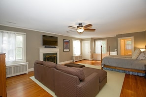 A spacious living room with a brown sectional sofa, a fireplace, a ceiling fan, and a large bed in the adjacent room. - 65 Blue Road: 6  BR, 6  BA House in Pinehurst, Sleeps 12 (Pinehurst)