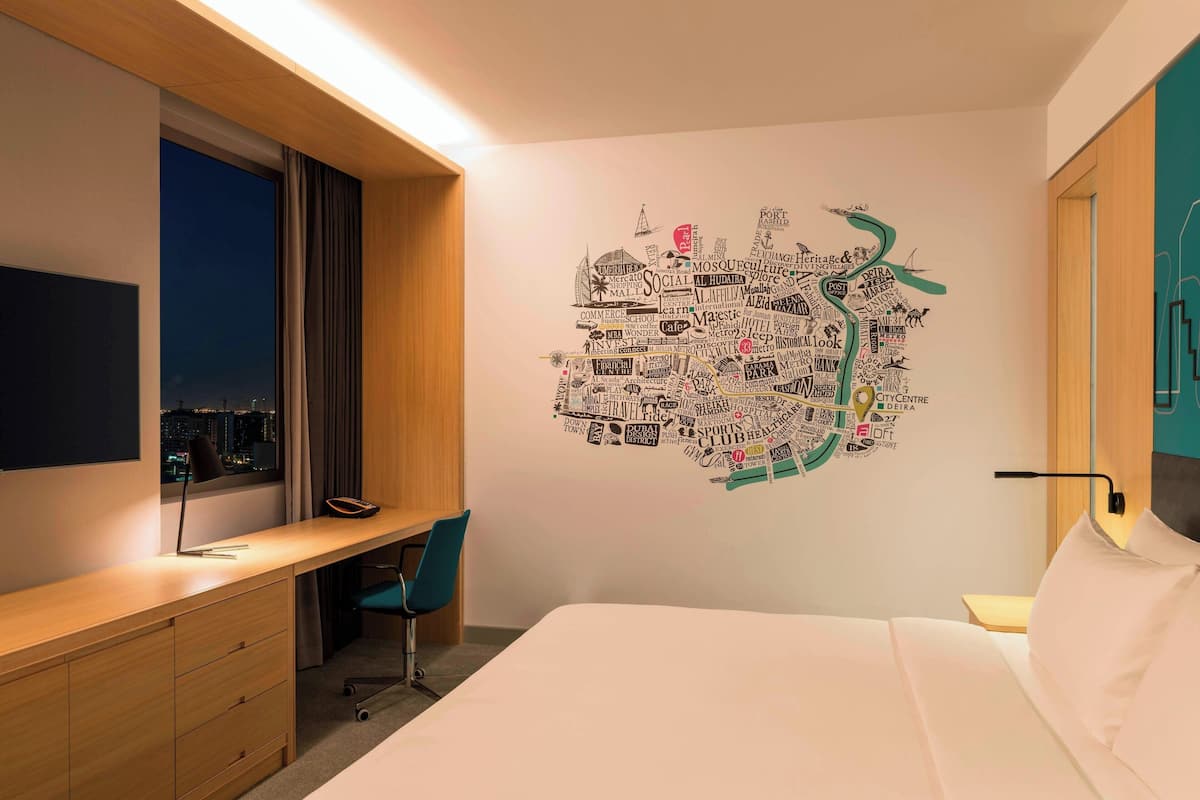 A hotel room with a large bed, a desk with a chair, a TV, and a wall art piece depicting a cityscape.