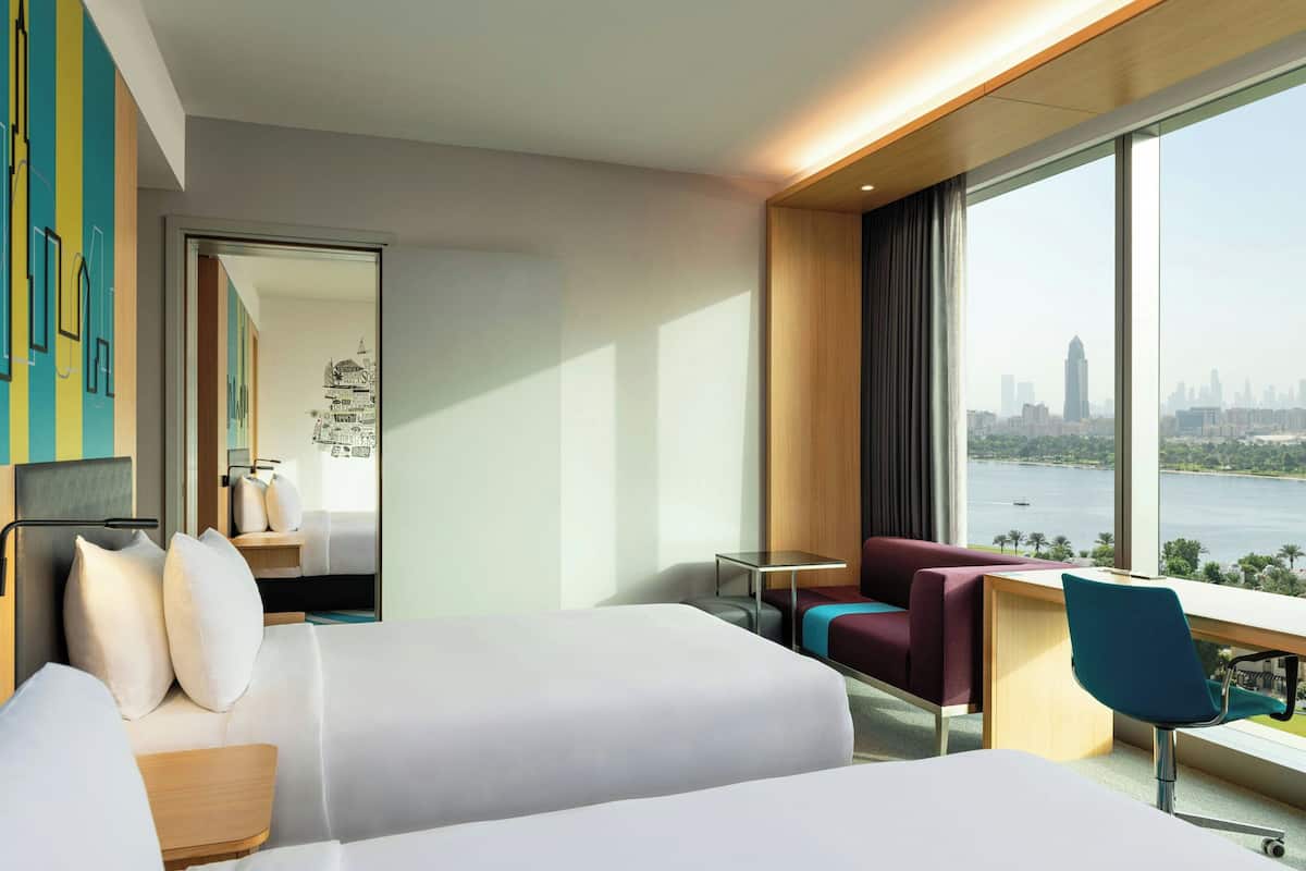 A modern hotel room with two beds, a desk, and a view of a city skyline and water.