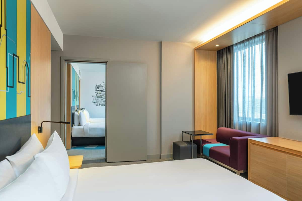 A modern hotel room with a large bed, a seating area with a sofa and armchair, a small table, and a TV mounted on the wall.