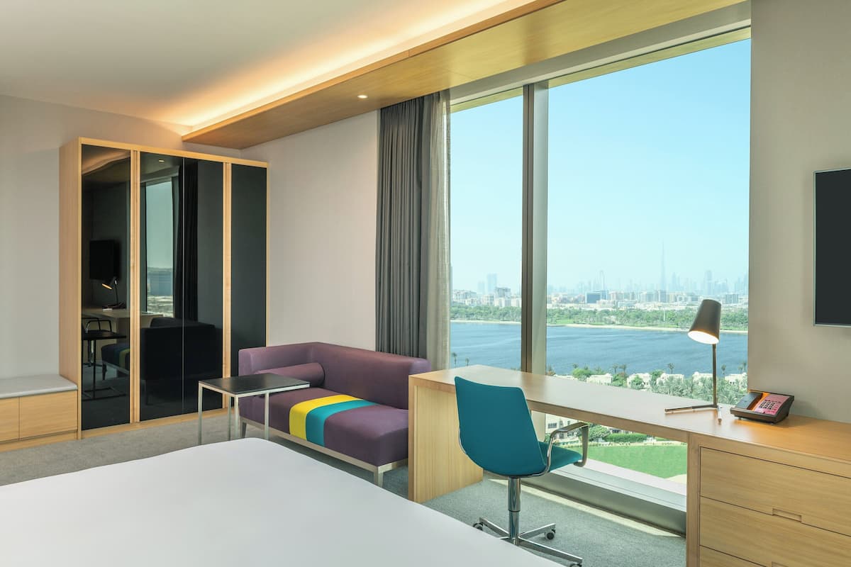 A hotel room with a bed, a desk, a chair, a sofa, a wardrobe, and a large window with a city view.