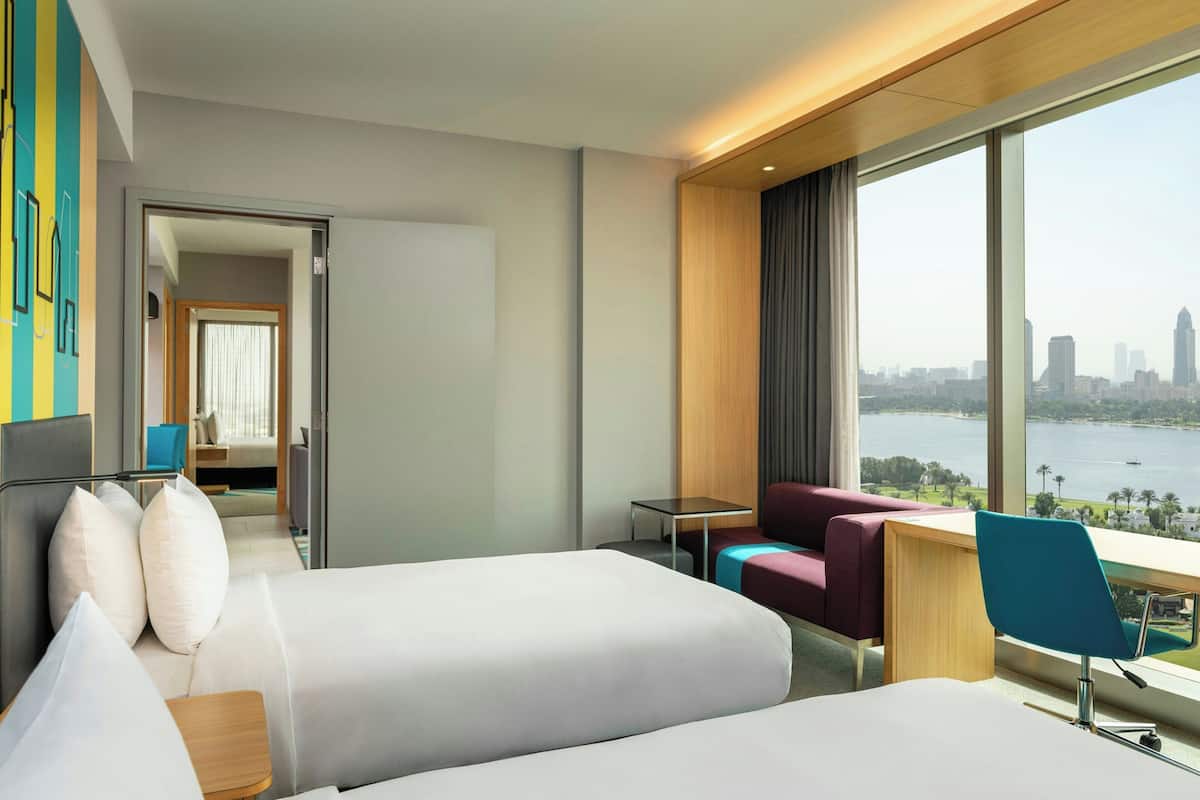A hotel room with two beds, a desk, a chair, a view of a city skyline and a body of water.