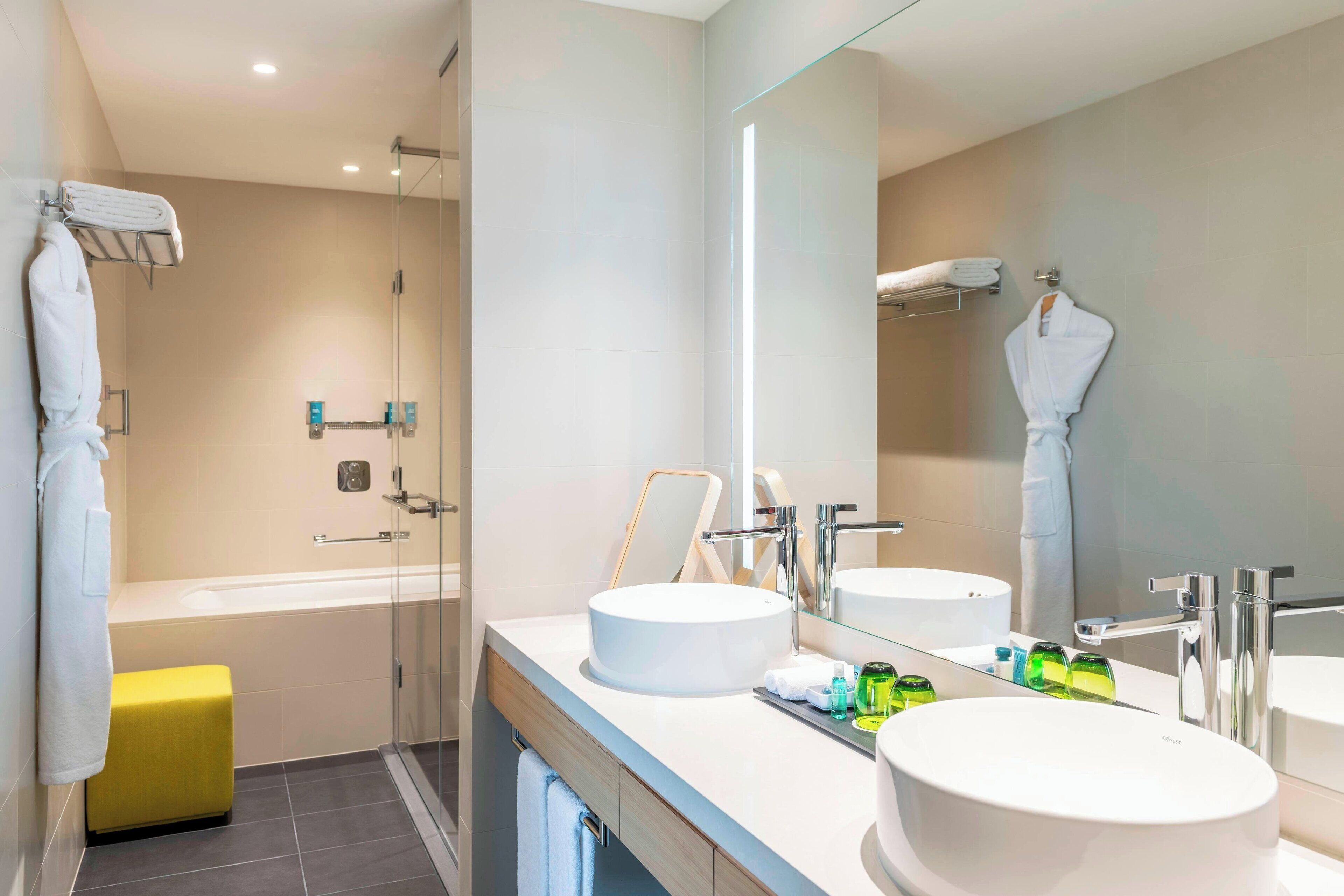 A modern bathroom with two sinks, a glass shower enclosure, and a yellow bench.