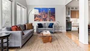 A modern living room with a grey sofa, a glass coffee table, and a cityscape painting on the wall. - Oasis in the City. Morningside/VaHi. Great in town location. (Atlanta)