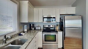 A kitchen with white cabinets, stainless steel appliances, granite countertops, and a window with blinds. - Hicks | Walk to the Beach + Pet Friendly! (Surf City)