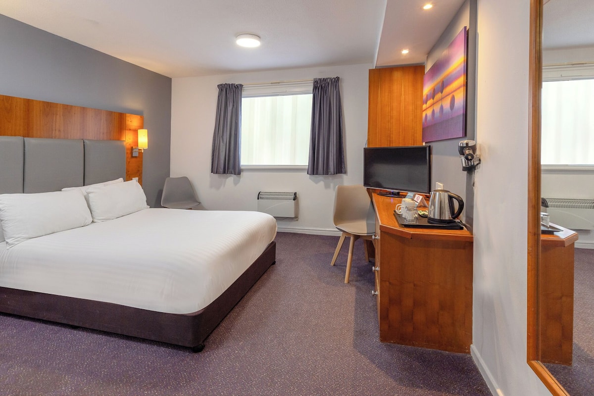 A hotel room with a large bed, a desk with a kettle, a chair, and a TV.