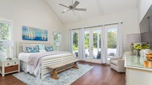 Une chambre à coucher comprenant un lit, une table de chevet, une commode, un ventilateur de plafond et une grande fenêtre avec des rideaux.