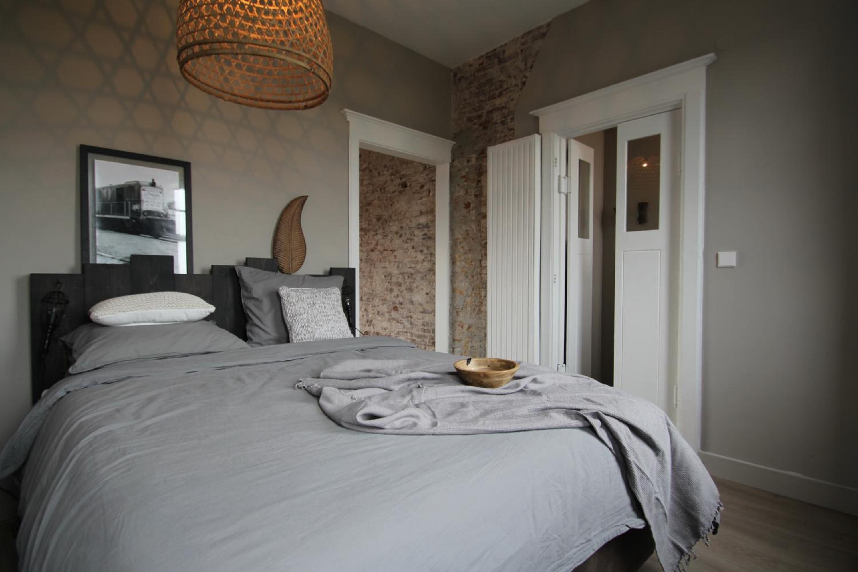 A bedroom with a large bed, a wooden bowl on the bed, a woven light fixture, and a brick wall.