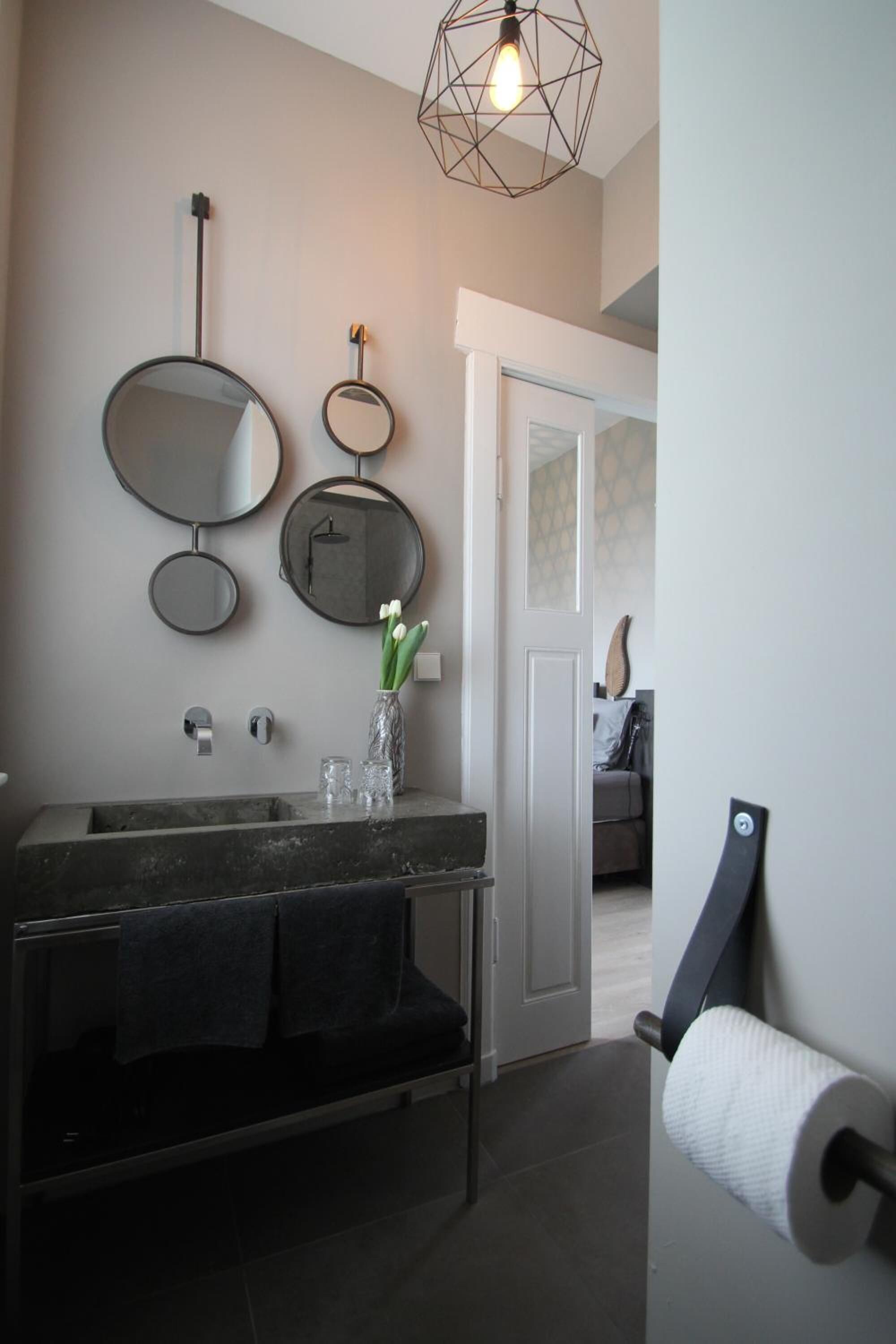 A modern bathroom with a dark granite sink, three wall-mounted mirrors, a vase with flowers, and a geometric light fixture.